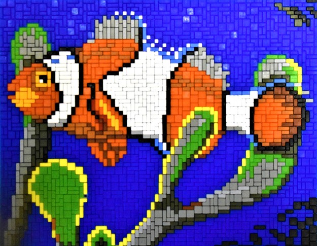 Mosaik-Pixelart eines Clownfischs in Orange und Weiß vor blauem Hintergrund, begleitet von grünen Seetang-Elementen